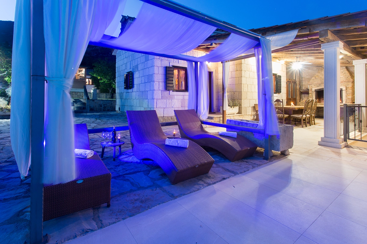 Modern, luxurious sun loungers in front of the villa, illuminated by the dark blue light of the pool at night, with the villa visible in the background.